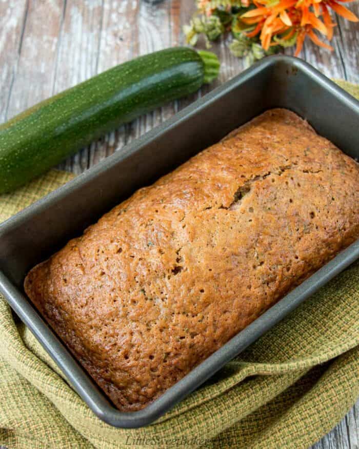 A zucchini loaf in a baking pan with a green dish towel and fresh zucchini beside it.