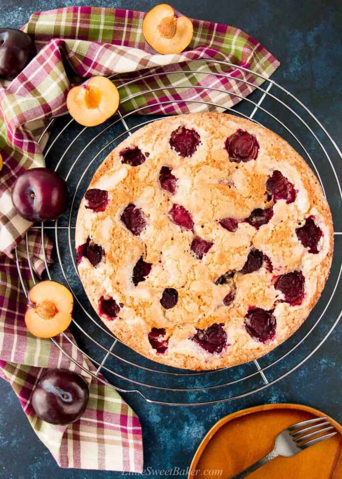 A fresh plum cake on a wire rack with a plaid dish towel and plums around it.