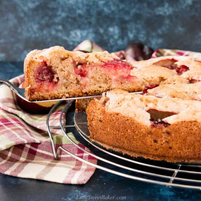 A slice of plum cake being removed from the cake on a wire rack.