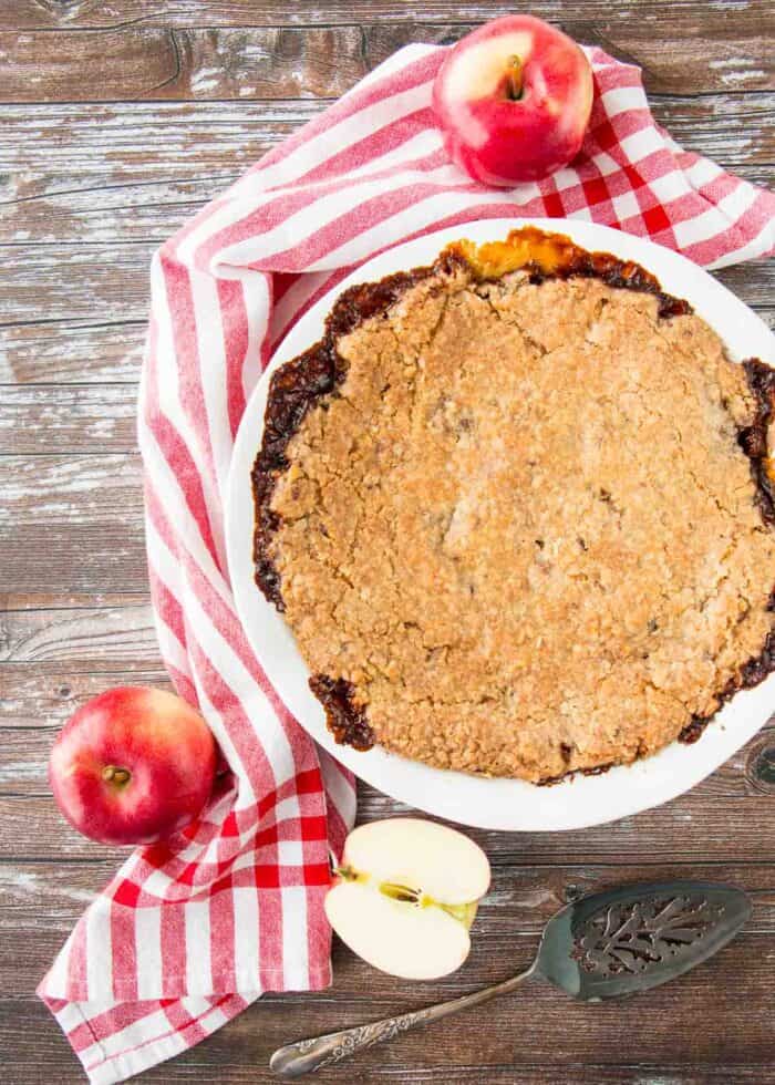 A whole Swedish apple oatmeal pie on a wooden table with a red striped dish clothe and apples around it.