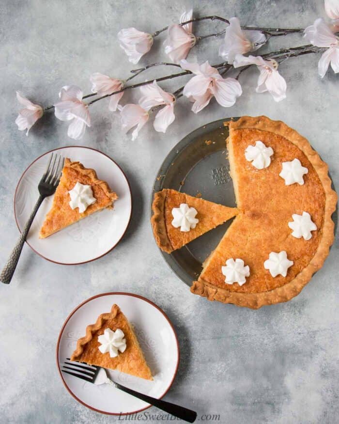 A chess pie in a pie pan with two slices plated and light pink flowers on a light gray background.