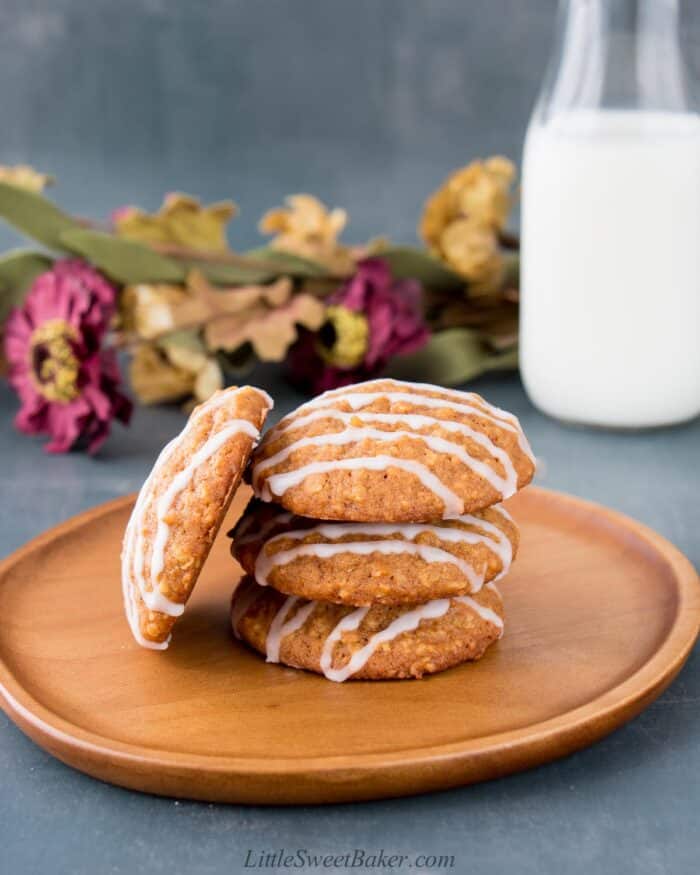 Four pumpkin oatmeal cookies on a wooden plate with a glass of milk and flowers behind.