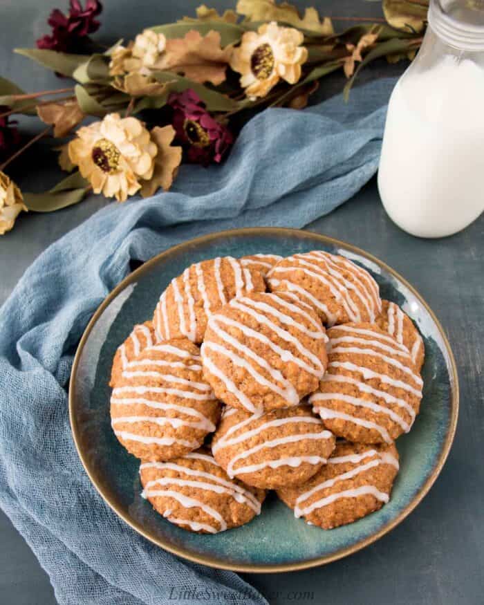 A plate of glazed pumpkin oatmeal cookies with a glass of milk, blue clothe and flowers.