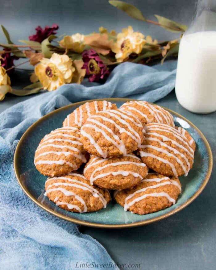 A plate of pumpkin oatmeal cookies with a glass of milk, blue silk cloth and flowers.