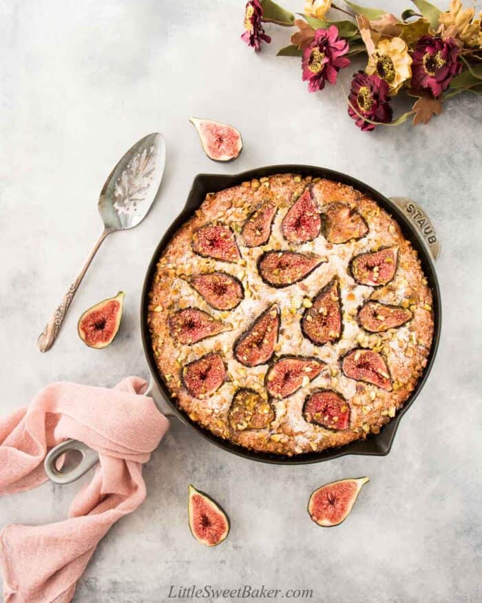 Fig cake in a skillet with some fresh figs around, pink cloth on handle and on a light gray backgorund.