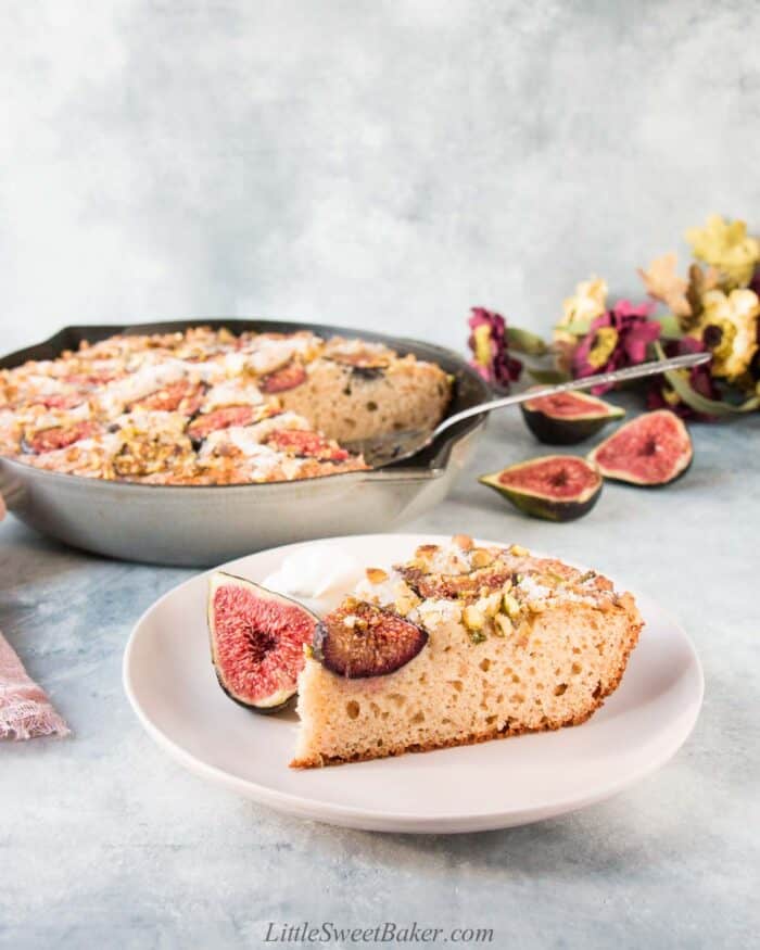 A slice of fig cake on a pale pink plate with the rest of the cake in a skillet in the background.