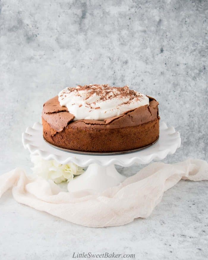 A flourless chocolate cake topped with whipped cream on a white cake stand.