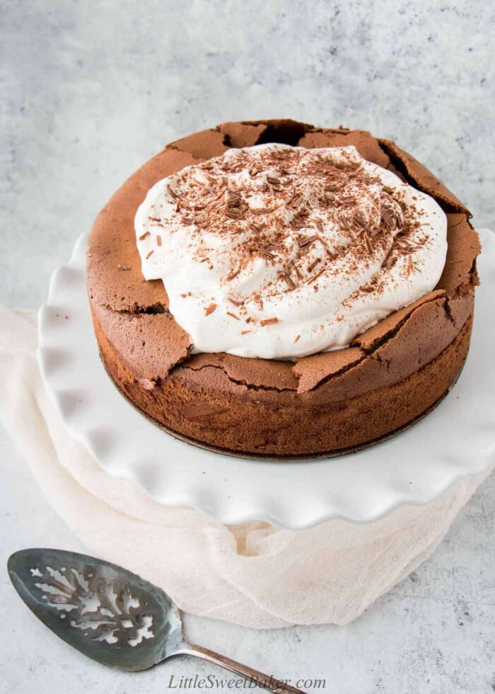 A flourless chocolate torte topped with whipped cream on a white cake stand.