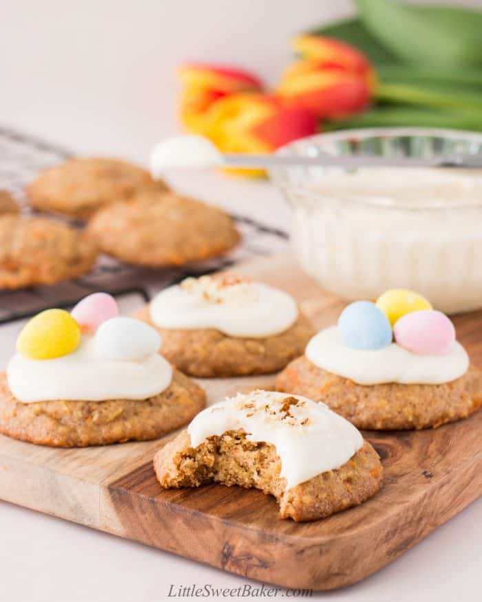 Carrot cake cookies with cream cheese frosting on a wooden board with a bite taken out of the front cookie.