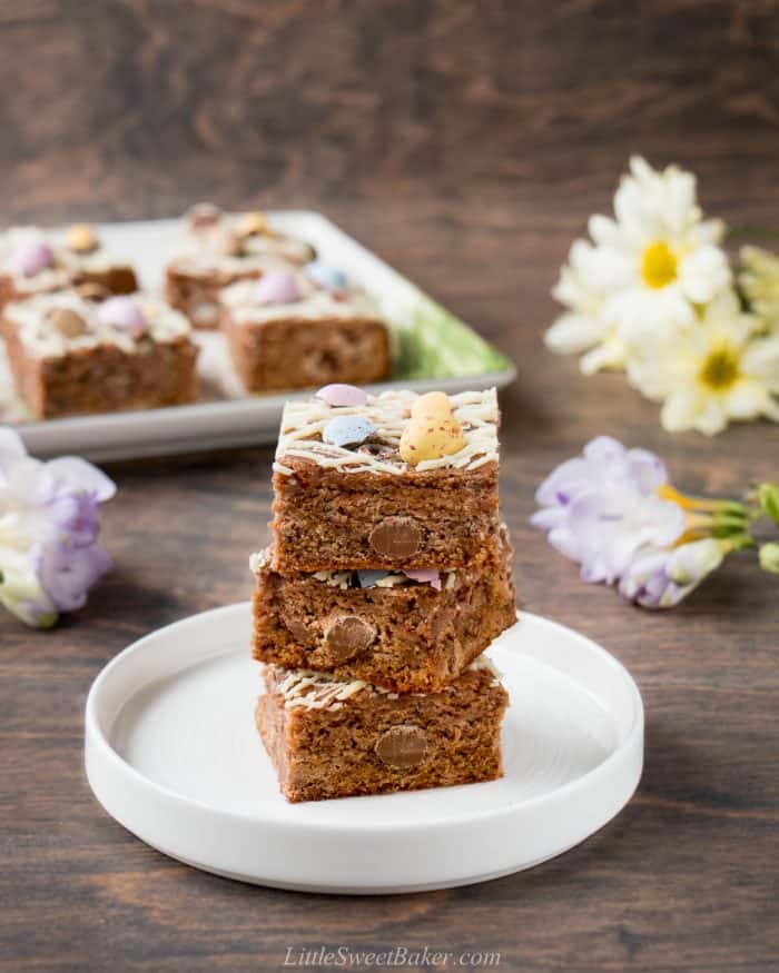 Malted milk blondies with mini eggs and white chocolate stacked on a white plate.