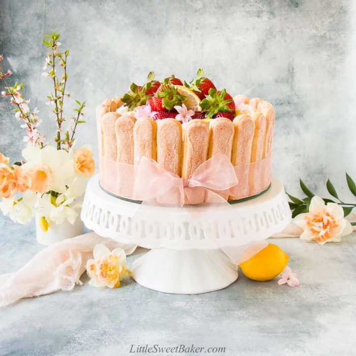 A strawberry lemon charlotte cake on a white cake stand surrounded with flowers.