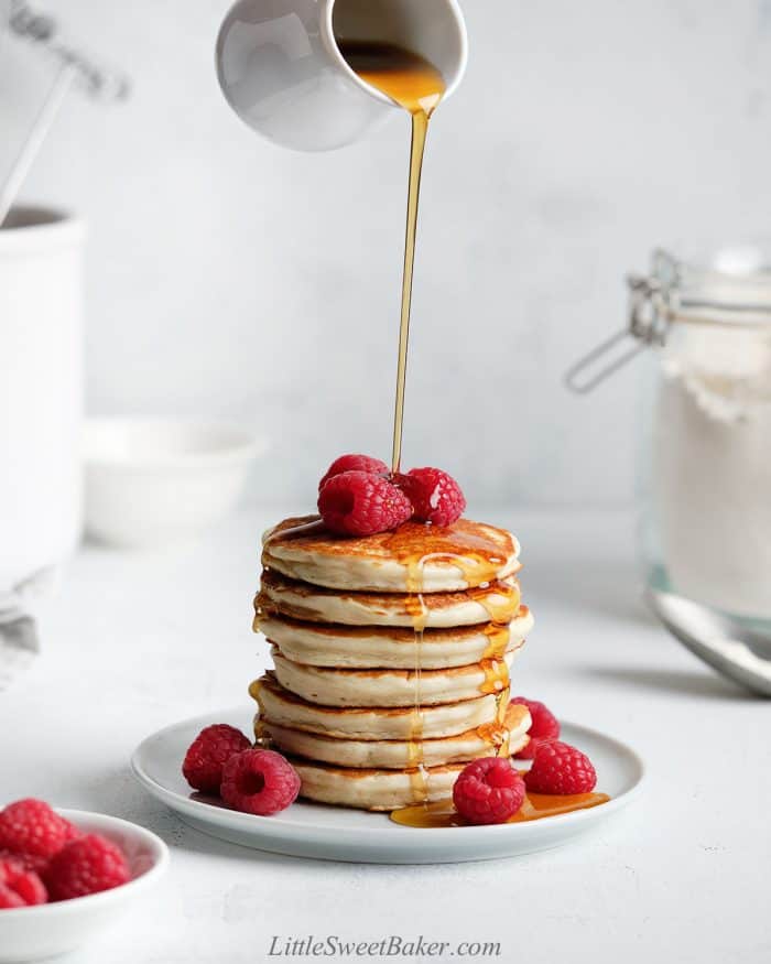 Maple syrup being poured over a stack of pancakes with fresh raspberries.