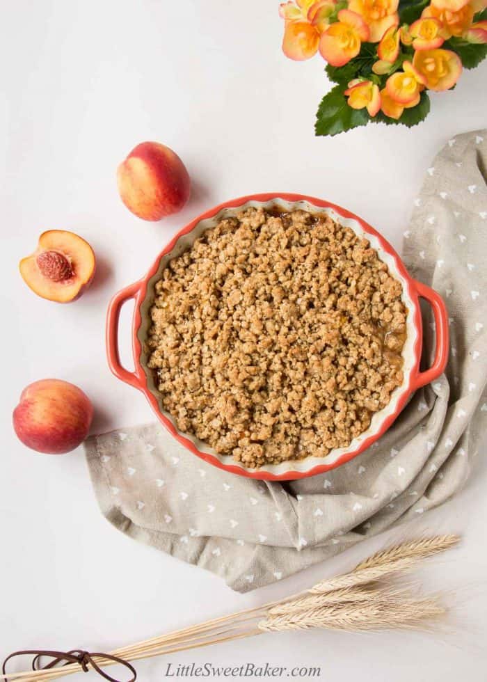 Peach crisp in a ceramic dish surrounded by peaches and beige napkin.