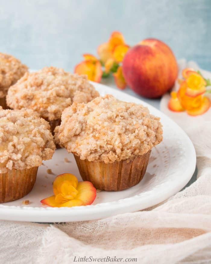 Peach crumb muffins on a white plate with a peach and flowers in the back.