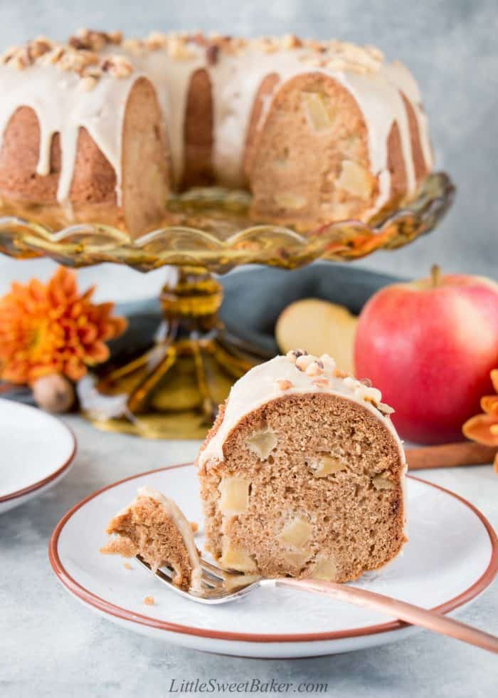 A slice of apple cake on a plate with a piece on a fork with the rest of the cake in the background.