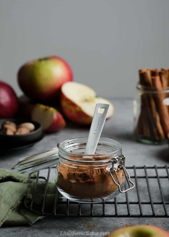 A jar of apple pie spice with a measuring spoon inside and apples and cinnamon sticks in the background.