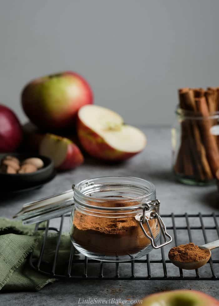 An open jar of apple pie spice on a cooling rack with apples and cinnamon sticks in the background.