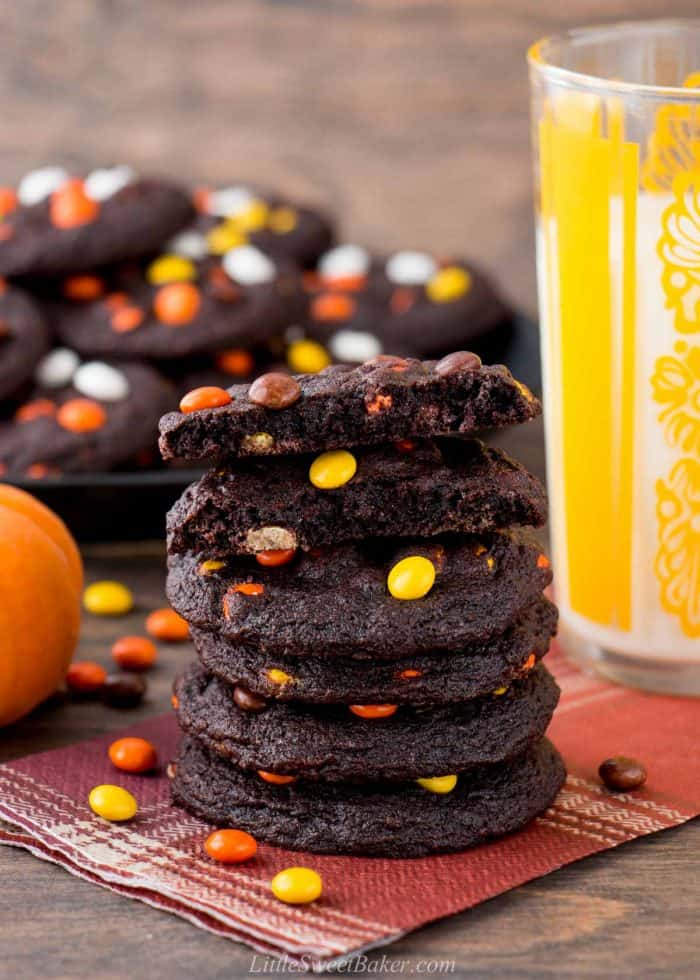 A stack of chocolate Reese's Pieces cookies on a napkin with a glass of milk.