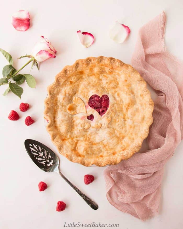 A whole raspberry pie with a pink cloth, rose, and raspberries scattered around it.