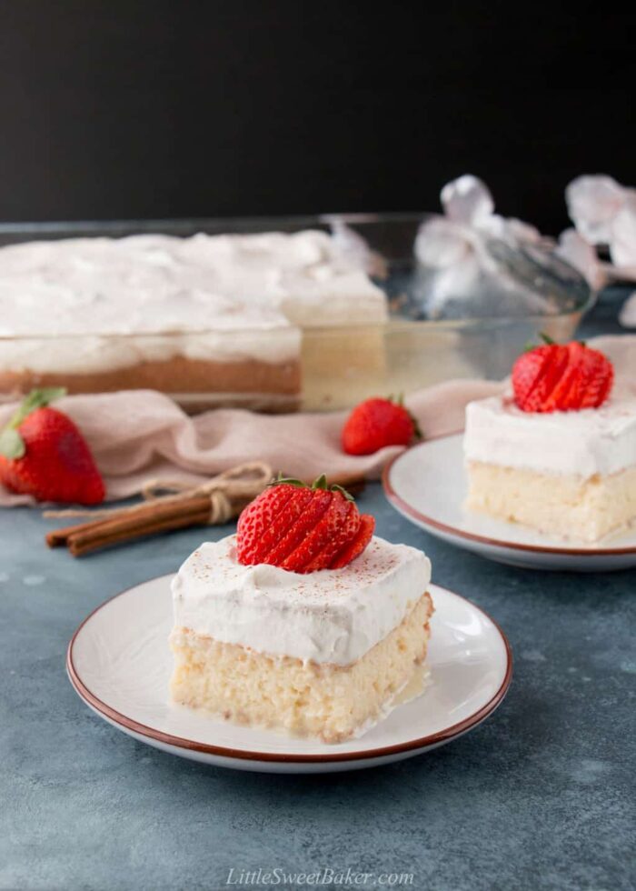 Two slices of tres leches cake topped with strawberries with the rest of the cake in the back.