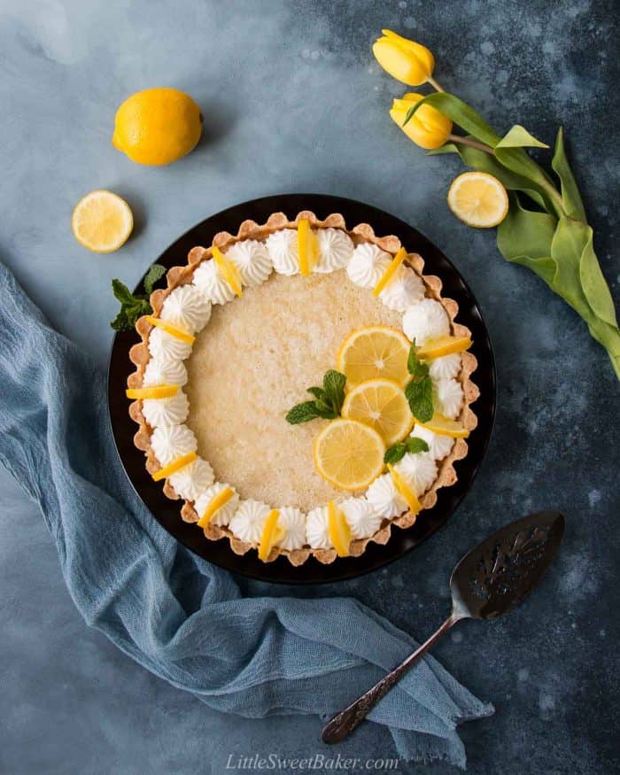 A whole lemon tart decorated with whipped cream and lemon slices on a black plate.
