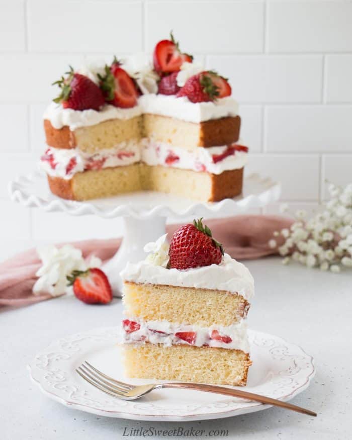 A slice of strawberry shortcake cake on a white plate with the rest of the cake in the back