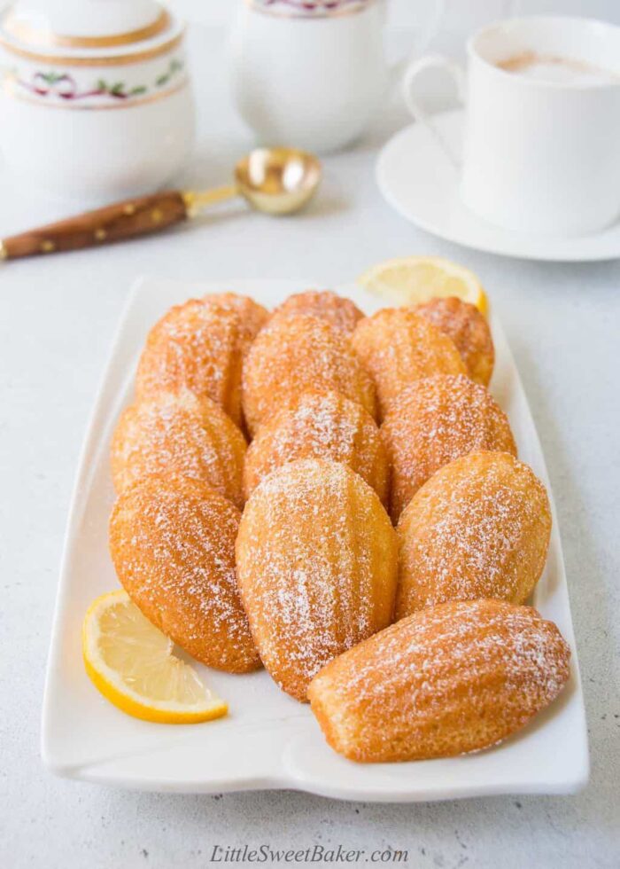 A white serving platter of madeleine cookies lightly dusted with powered sugar and lemon wedges.