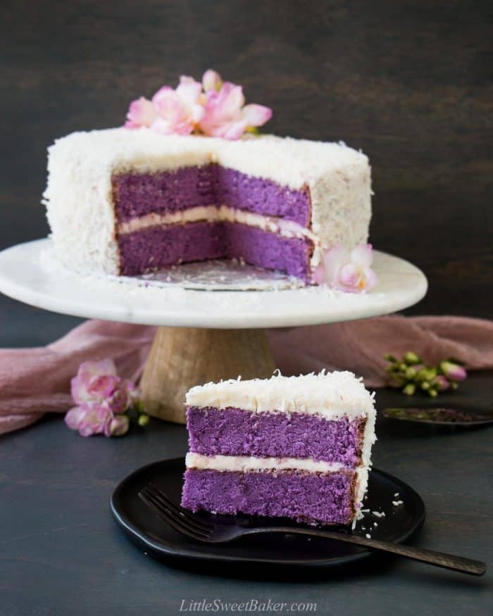 A slice of ube cake on a black plate with the rest of the cake in the back.