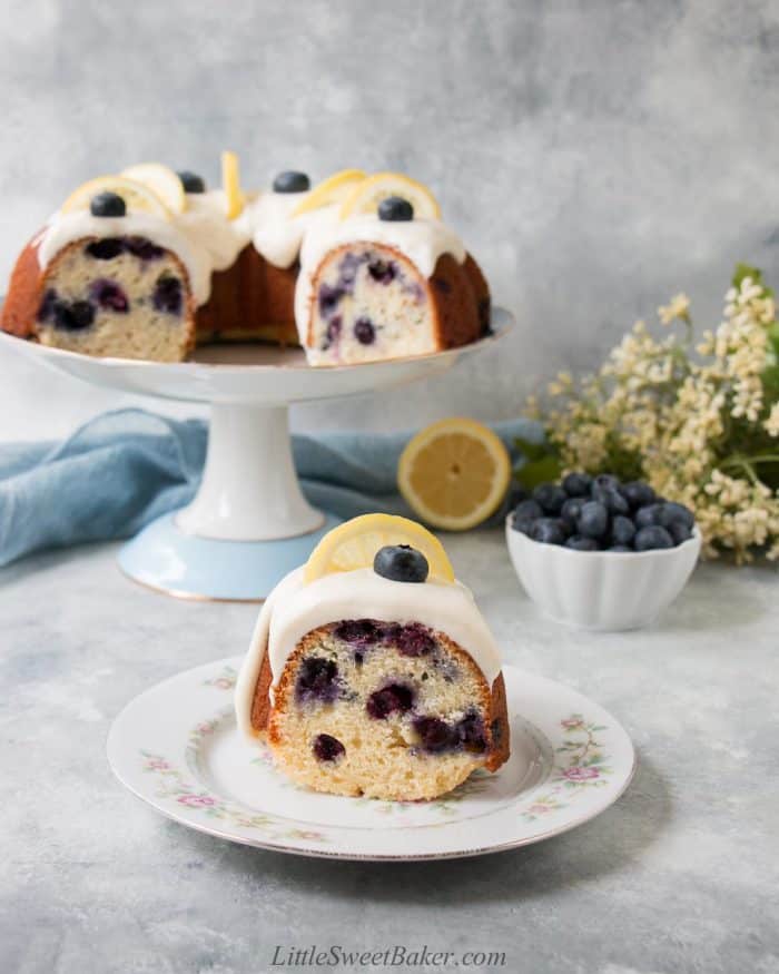 A slice of lemon blueberry cake on a vintage plate with the rest of the cake on a cake stand.