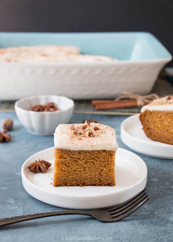 A slice of pumpkin cake topped with cream cheese frosting and pecans on a white plate.