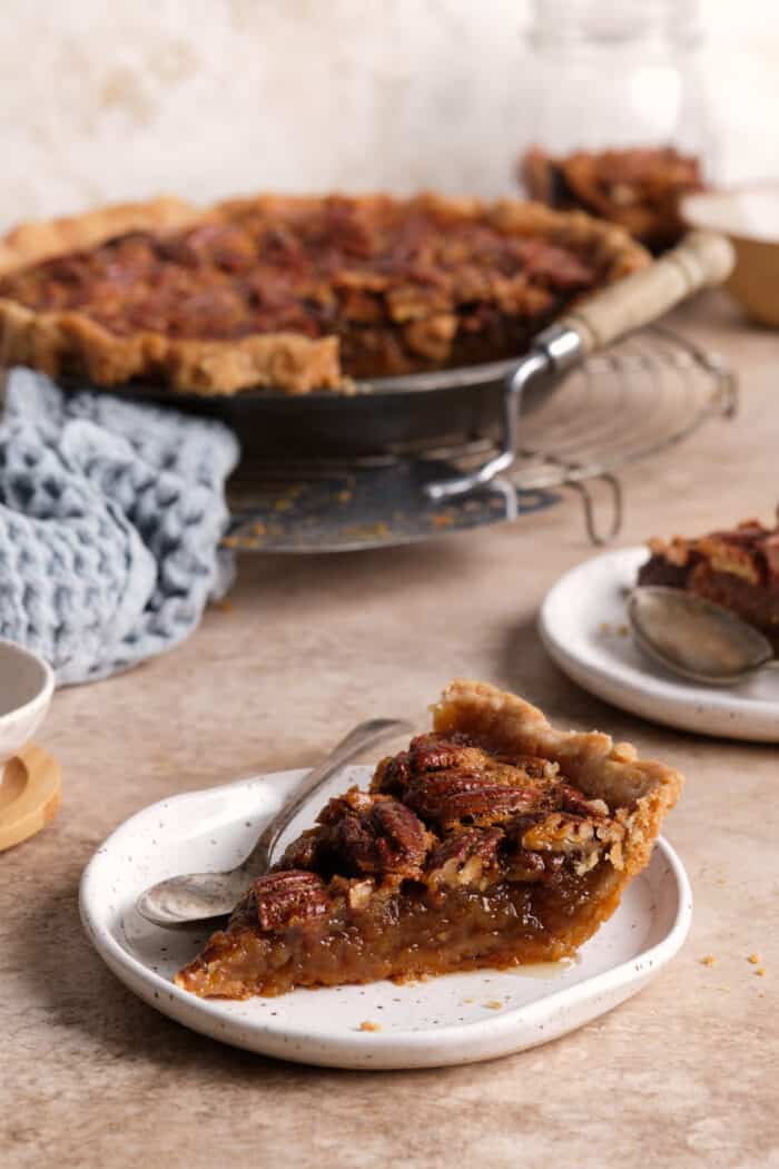 A slice of pecan pie on a white plate with the rest of the pie in the background