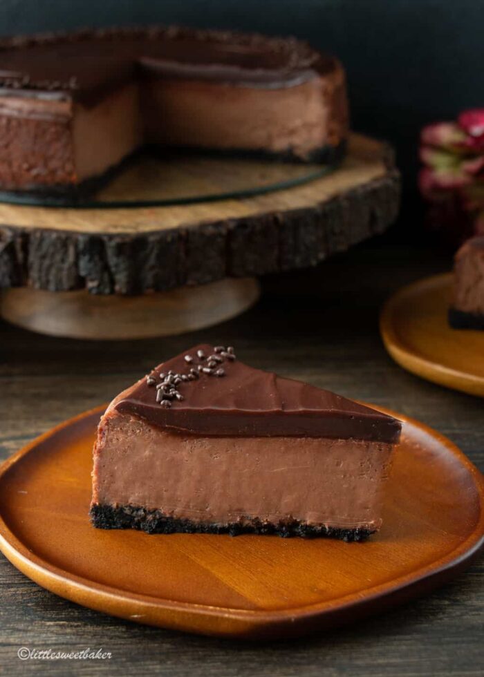 A slice of chocolate cheesecake topped with ganache on a wooden plate.