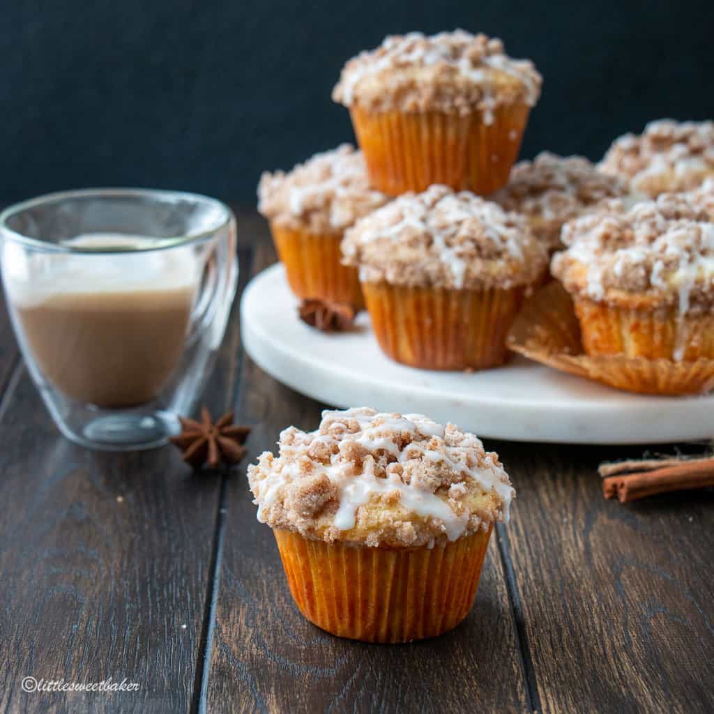 Coffee cake muffin on a wooden table with a cappuccino and more muffins in the background.