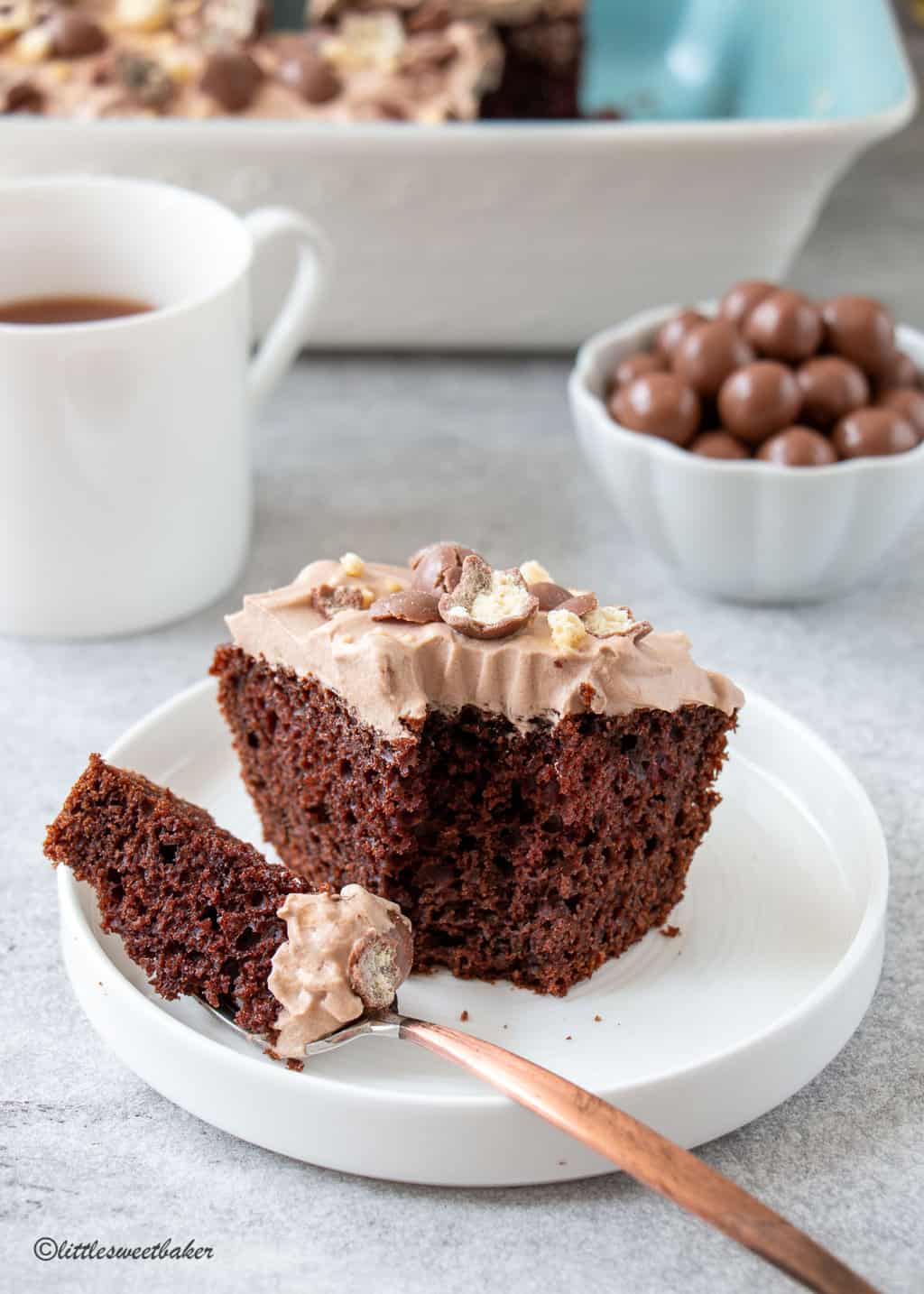 A slice of chocolate malt cake on a white plate with a piece on a fork.