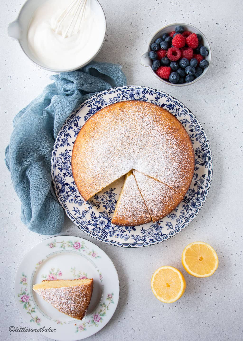 A lemon olive oil cake on a blue and white plate surrounded by a lemon, bowl of whipped cream, and blue cloth.