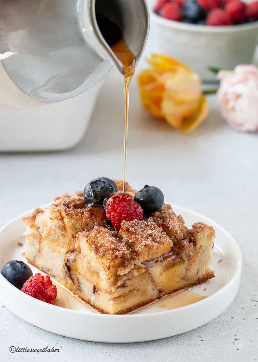 A slice of french toast casserole topped with a few berries and maple syrup being poured on top.