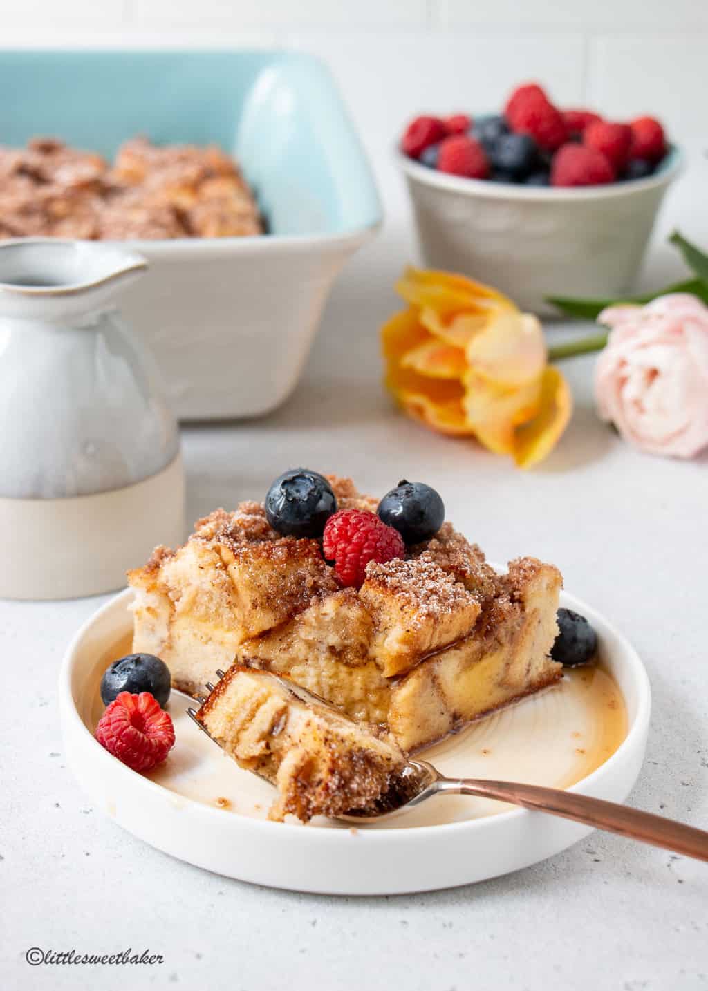 A slice of french toast casserole with berries and maple syrup on a white plate.