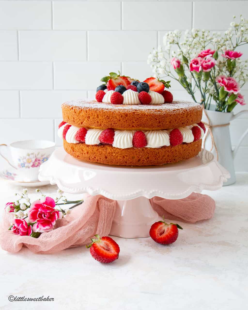 Victoria sponge cake topped with berries and cream on a pink cake stand.