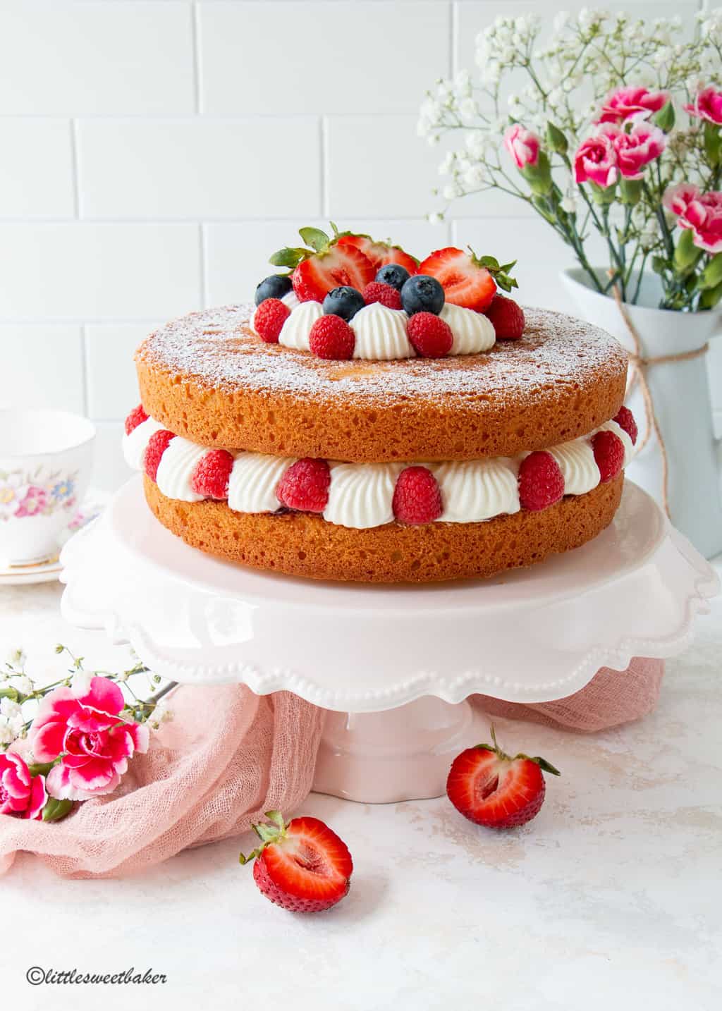 Victoria sponge cake topped with berries and cream on a pink cake stand.