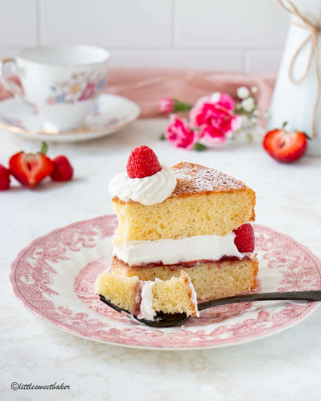 A slice of Victoria sponge cake on a pink plate with a piece on a fork.