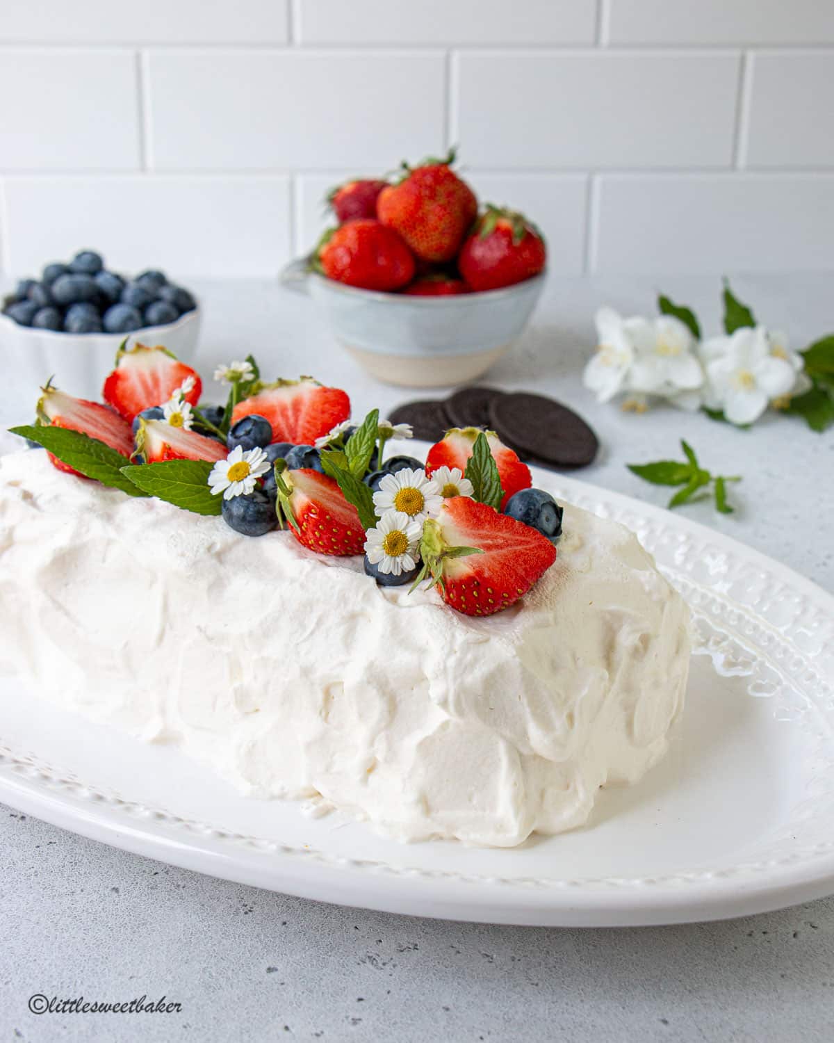 An icebox cake on a white plate topped with strawberries, blueberries, white flowers, and mint.