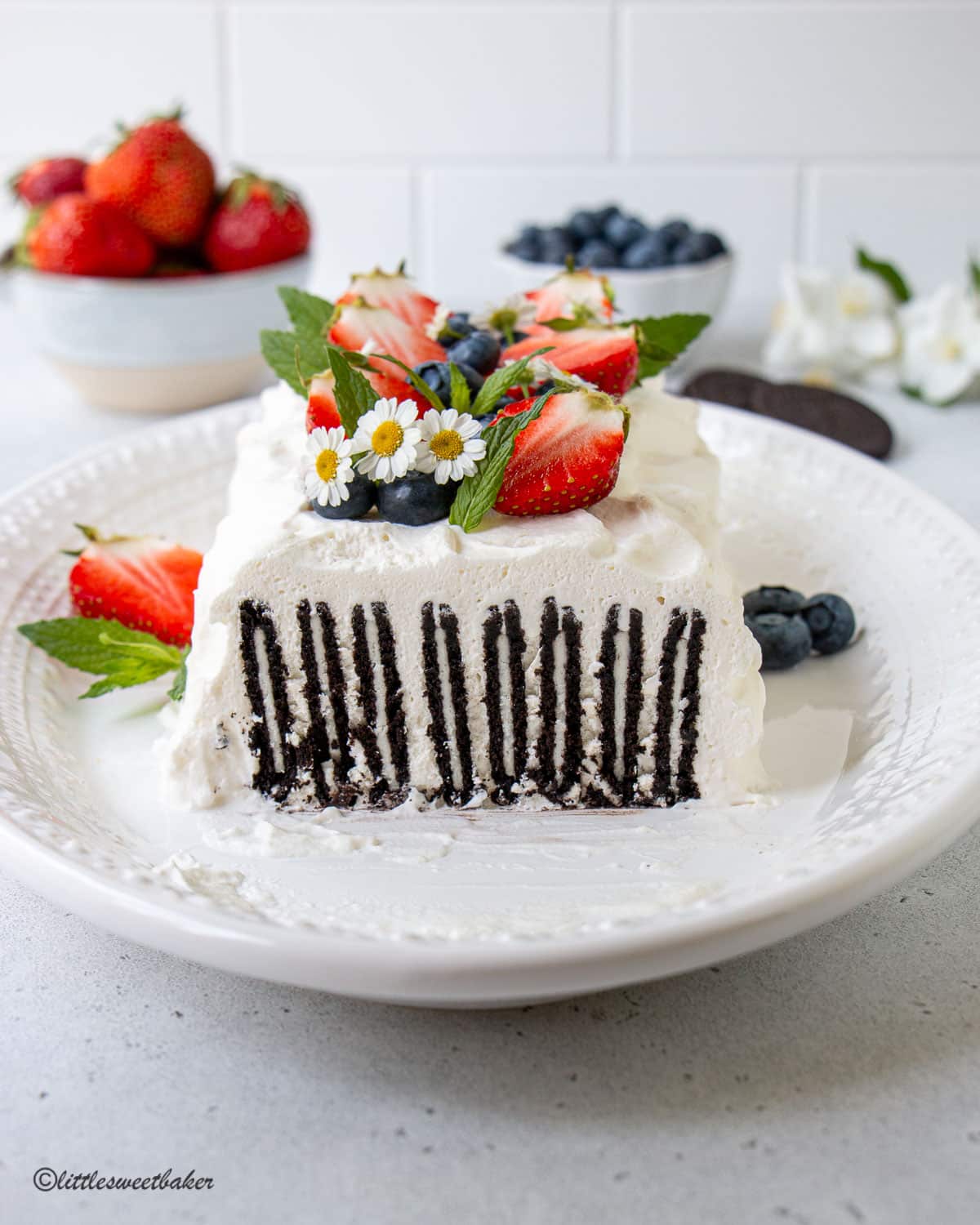 An icebox cake made with Oreos on a white plate topped with strawberries, blueberries, white flowers, and mint.
