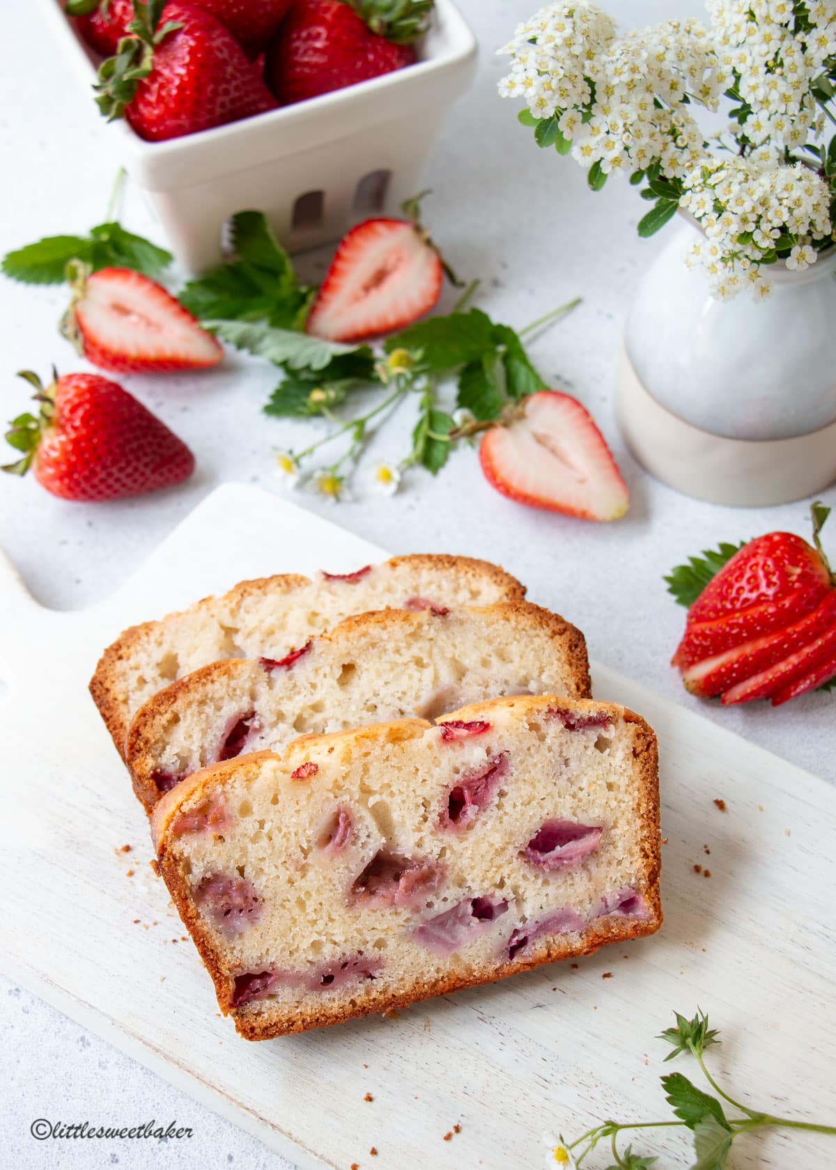 Three slices of strawberry bread on a white board with fresh strawberries scattered around.