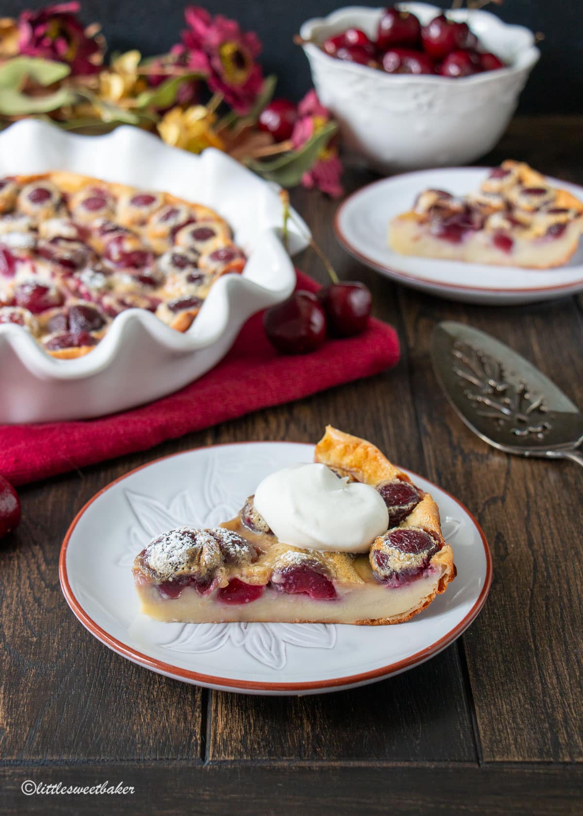 A slice of cherry clafoutis topped with whipped cream on a light grey plate.
