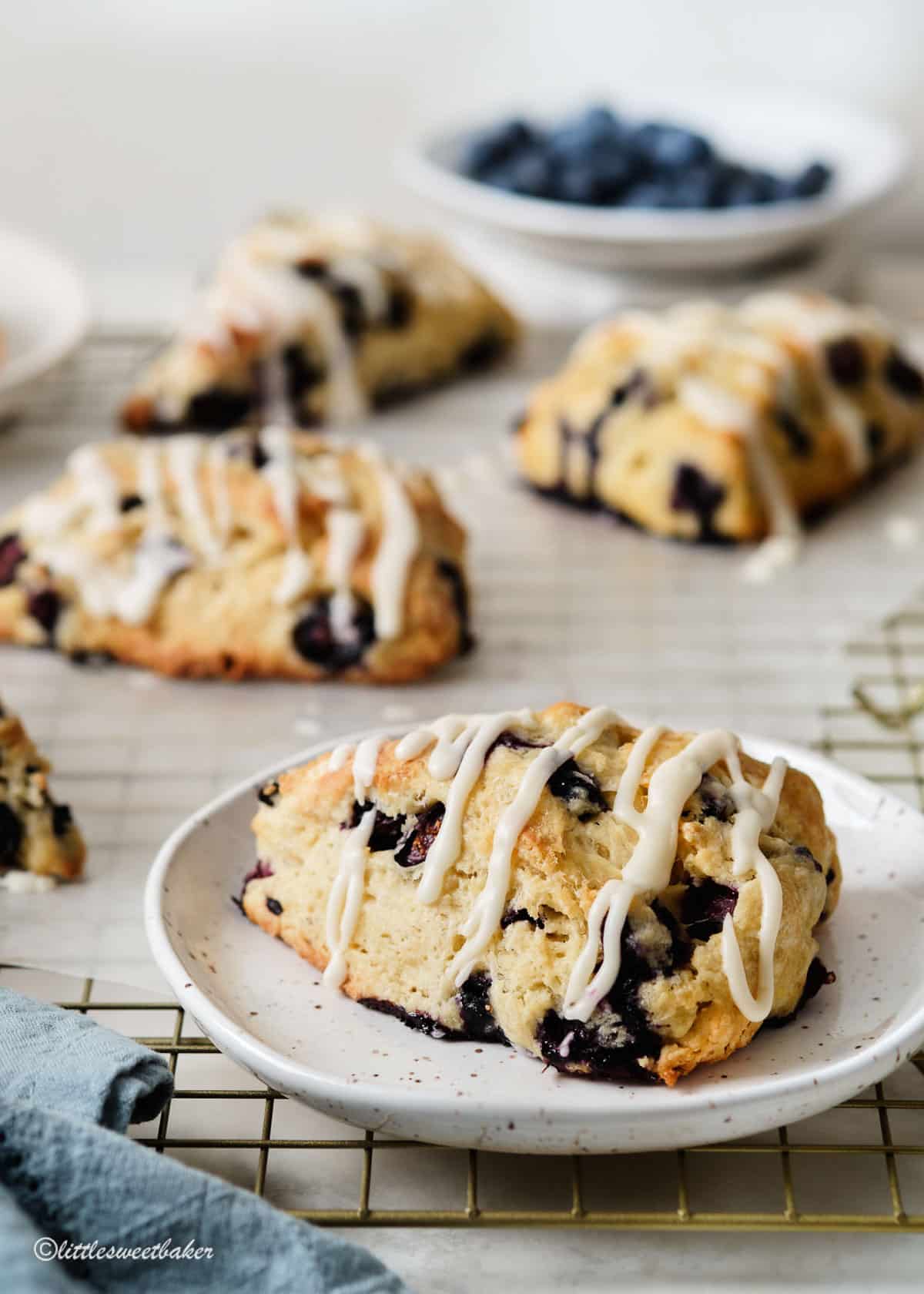 A glazed blueberry scone on a plate with other scones on a cooling rack.