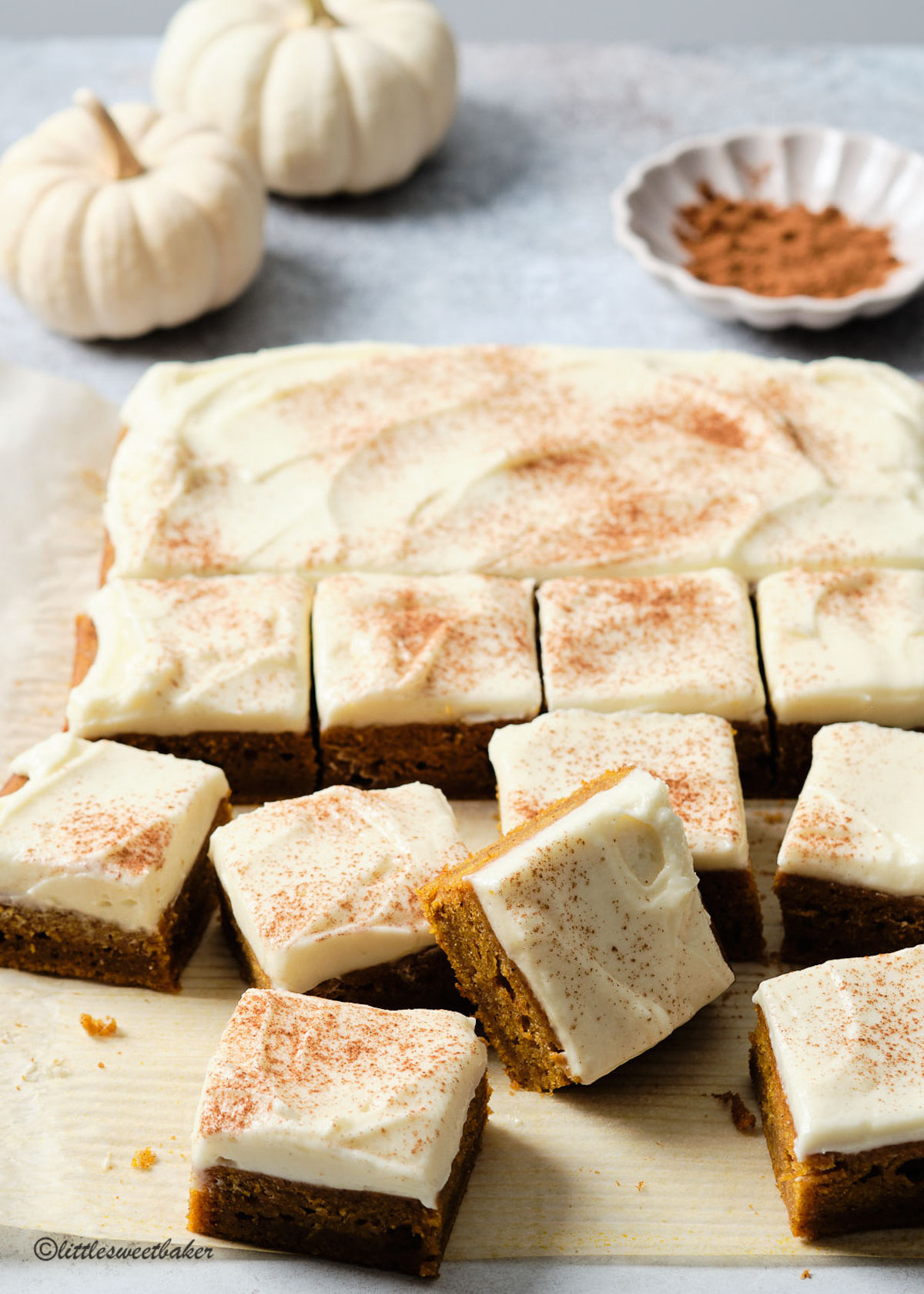 pumpkin bars half cut on parchment with white pumpkins and a small bowl of cinnamon in the background