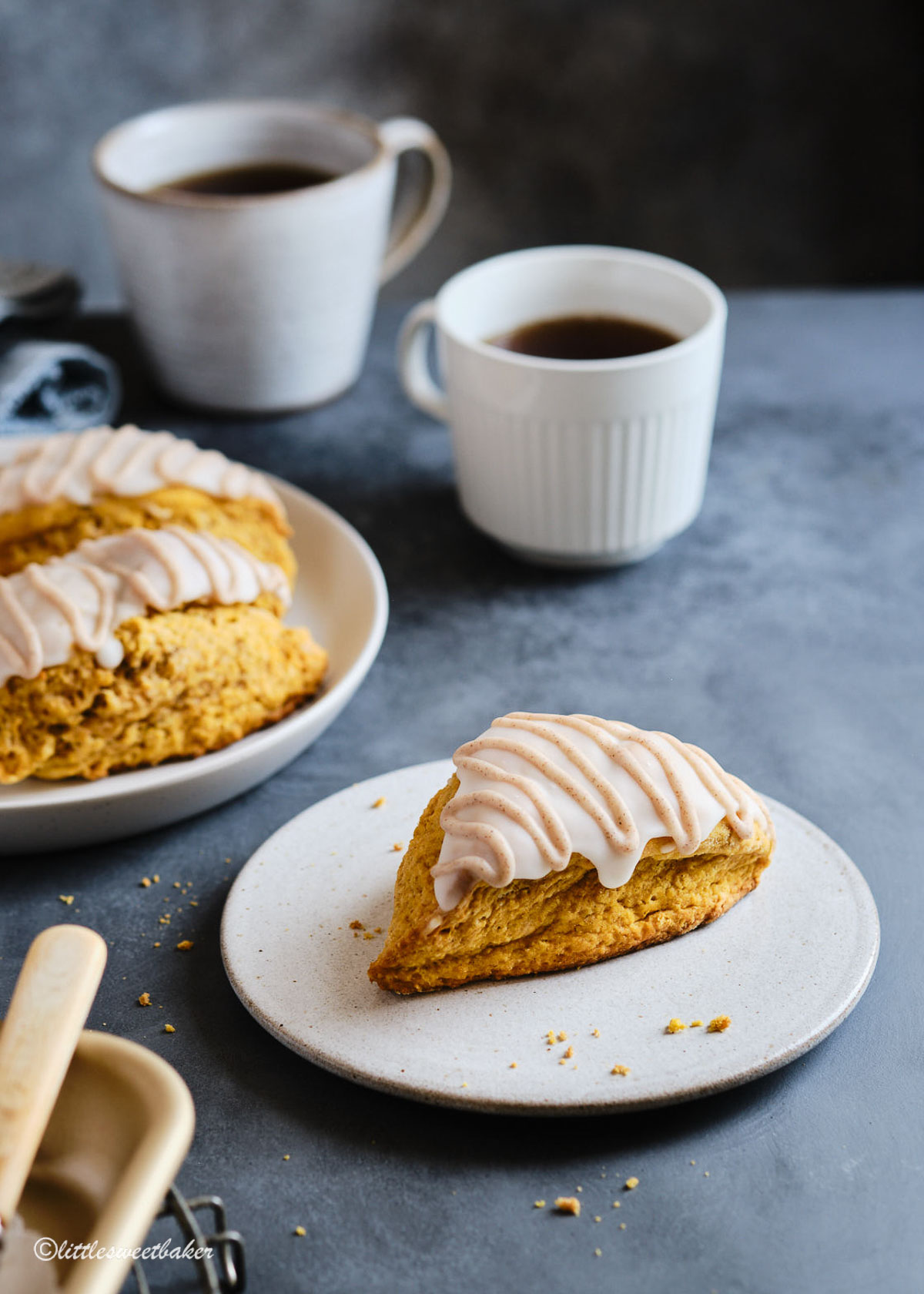 Glazed pumpkin scone on a plate with more in the back along with two cups of coffee.
