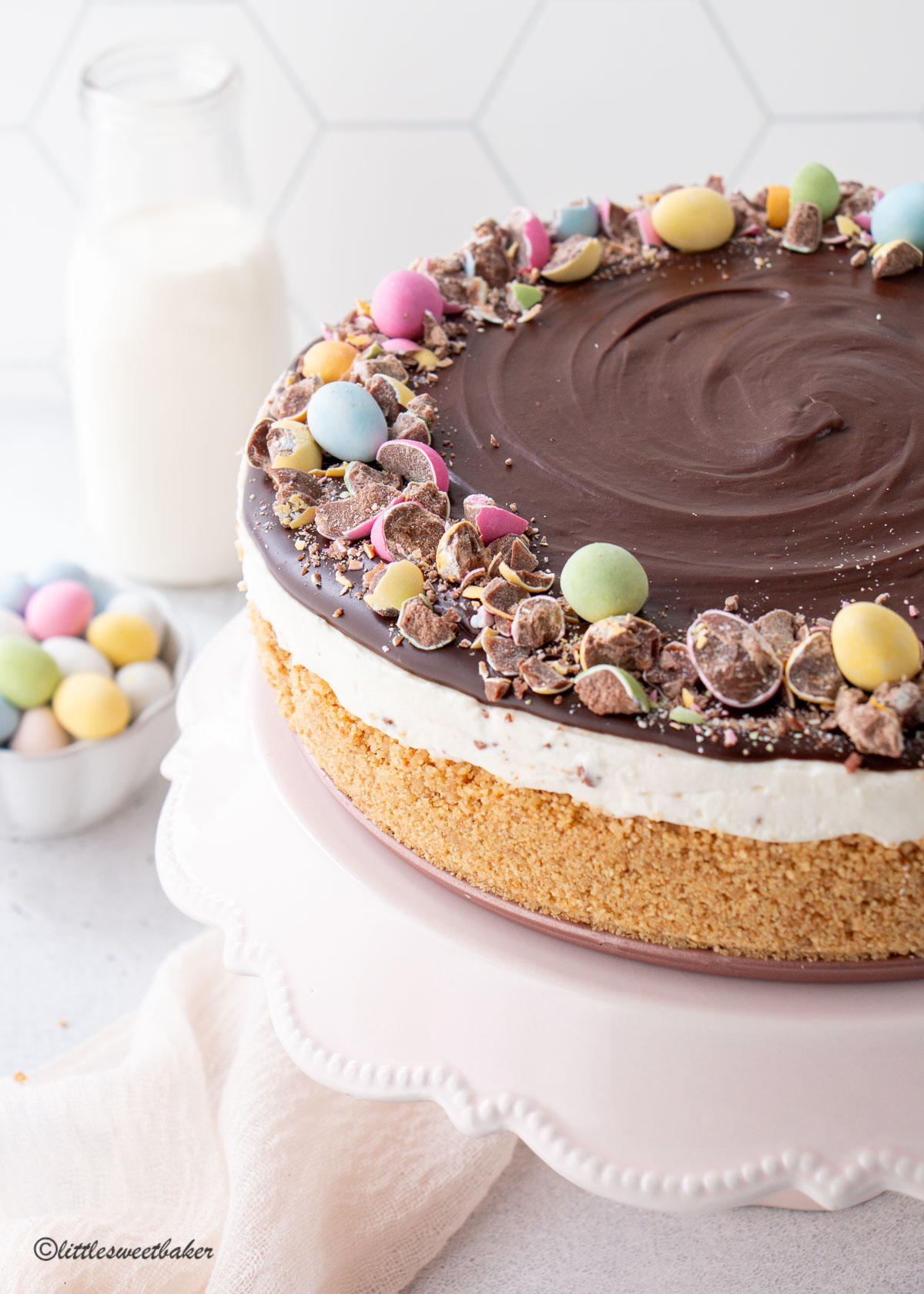 a mini egg cheesecake topped with chocolate ganache and crush mini eggs on a pink cake stand
