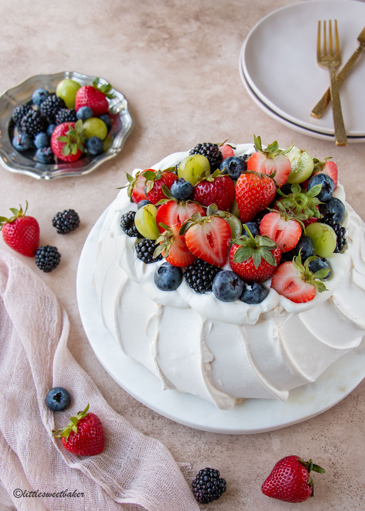A pavlova topped with fruit and whipped cream on a marble plate with a pink linen napkin and scattered fruits.