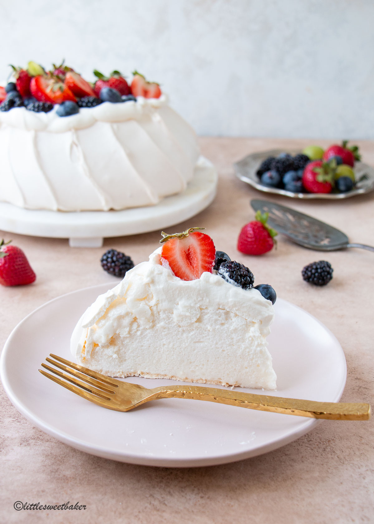 A slice of pavlova on a pink plate with a gold fork.